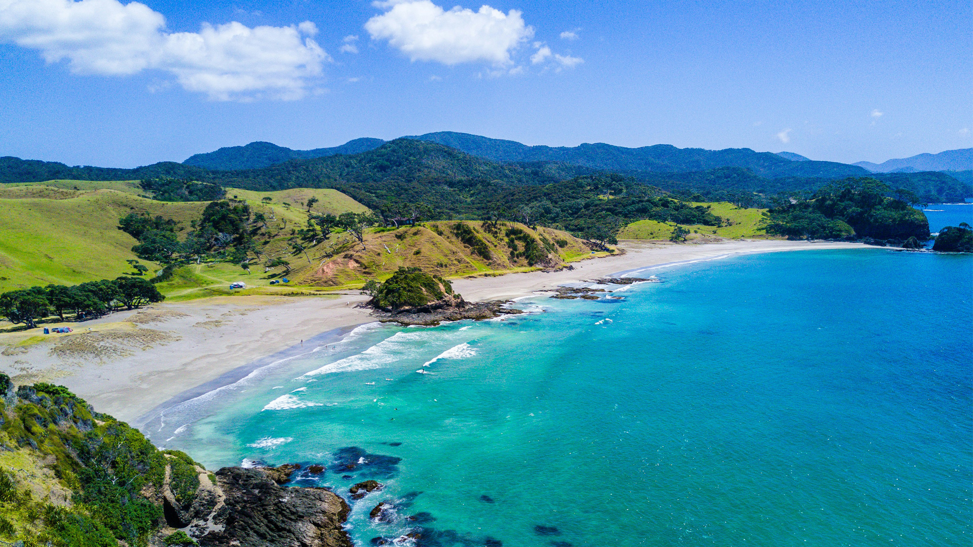 Aerial view of a curved white sandy beach with turquoise water, surrounded by green hills and trees under a partly cloudy blue sky.