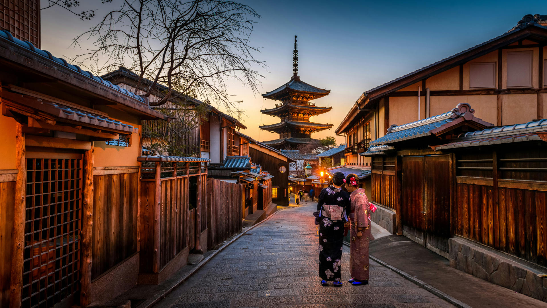 Two women dressed in traditional kimonos walking down a historic street with wooden buildings and a pagoda in the background at sunset in Japan.