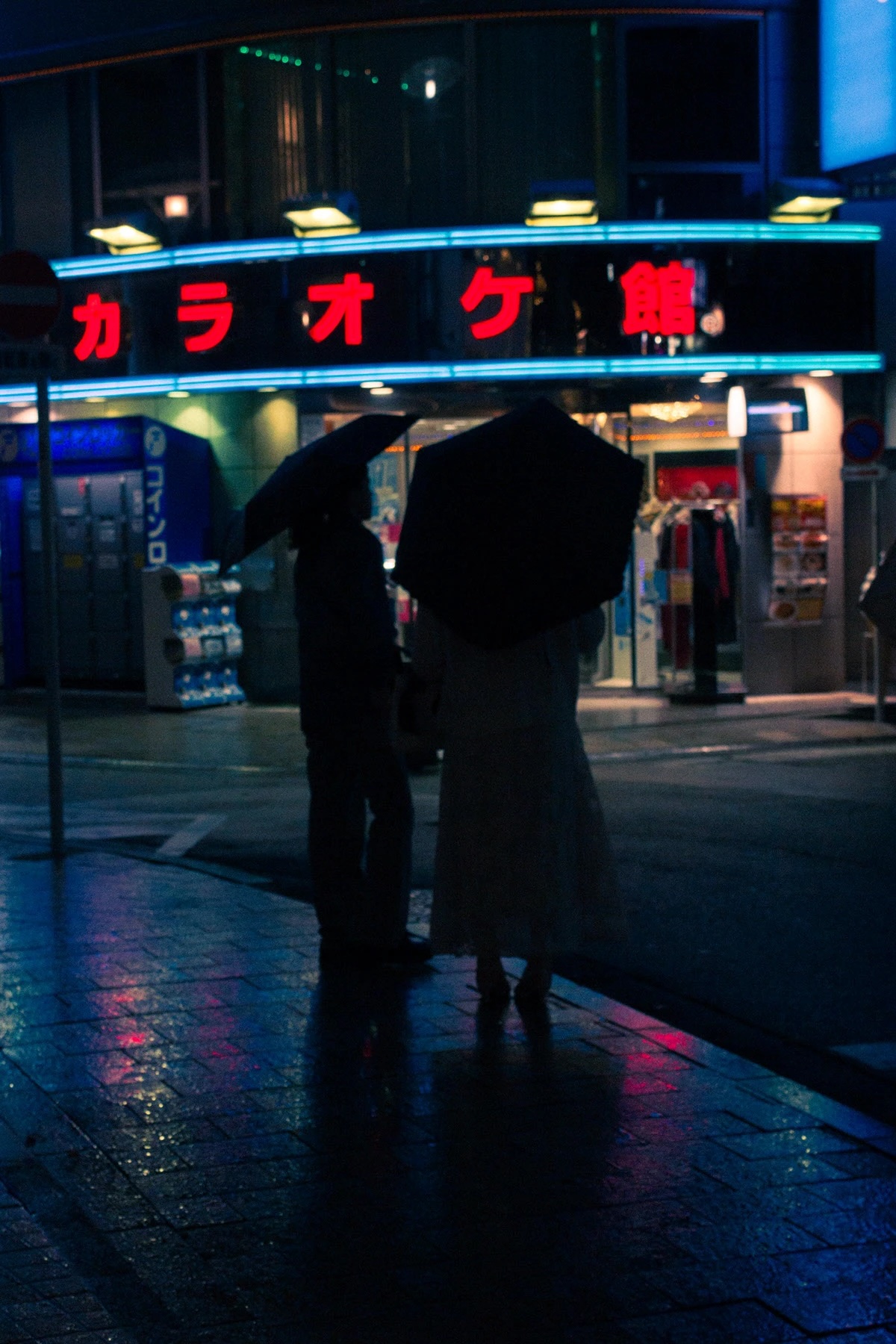 Two people holding umbrellas walking on a wet city street at night with neon Japanese karaoke sign in the background.