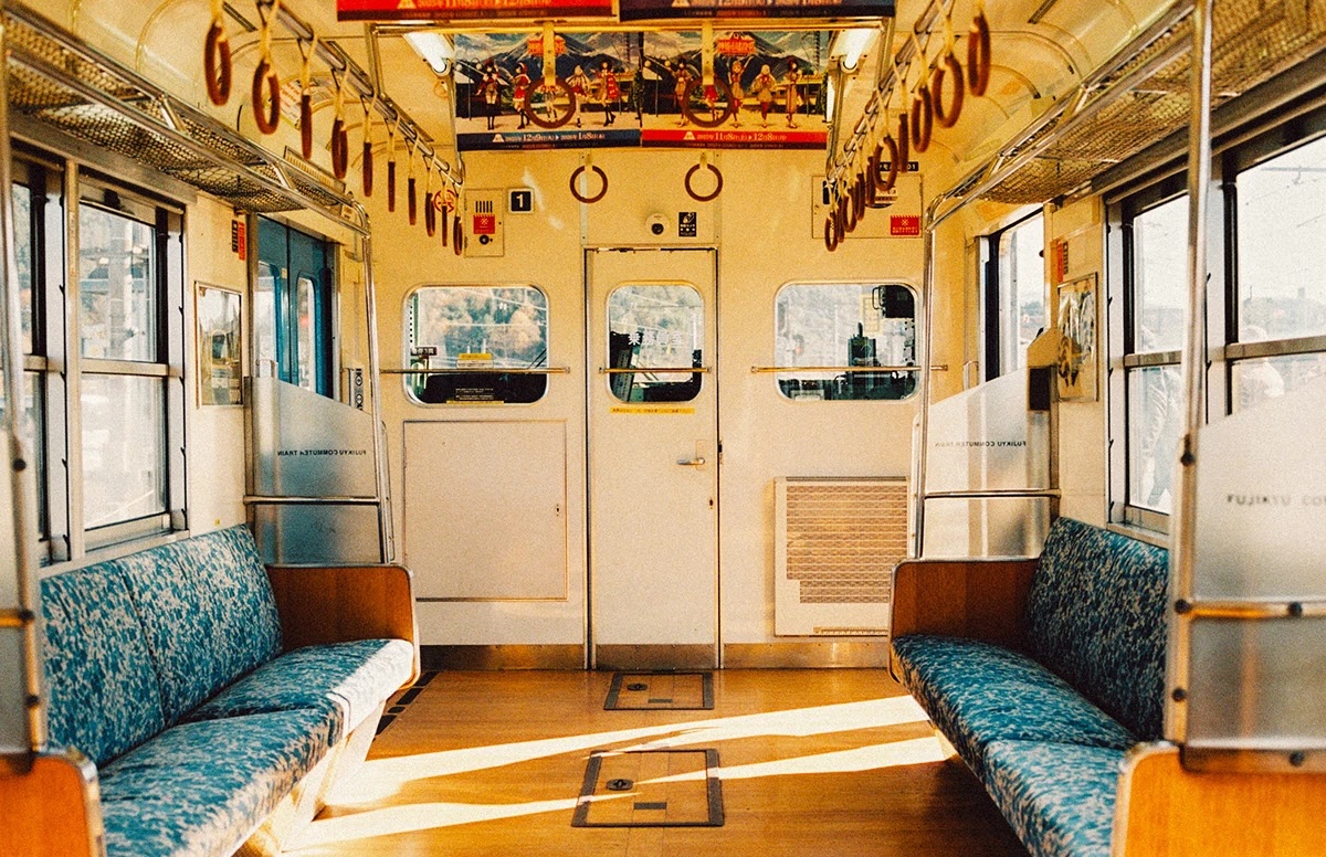 Empty interior of a sunlit train car with patterned blue seats and hanging hand straps.