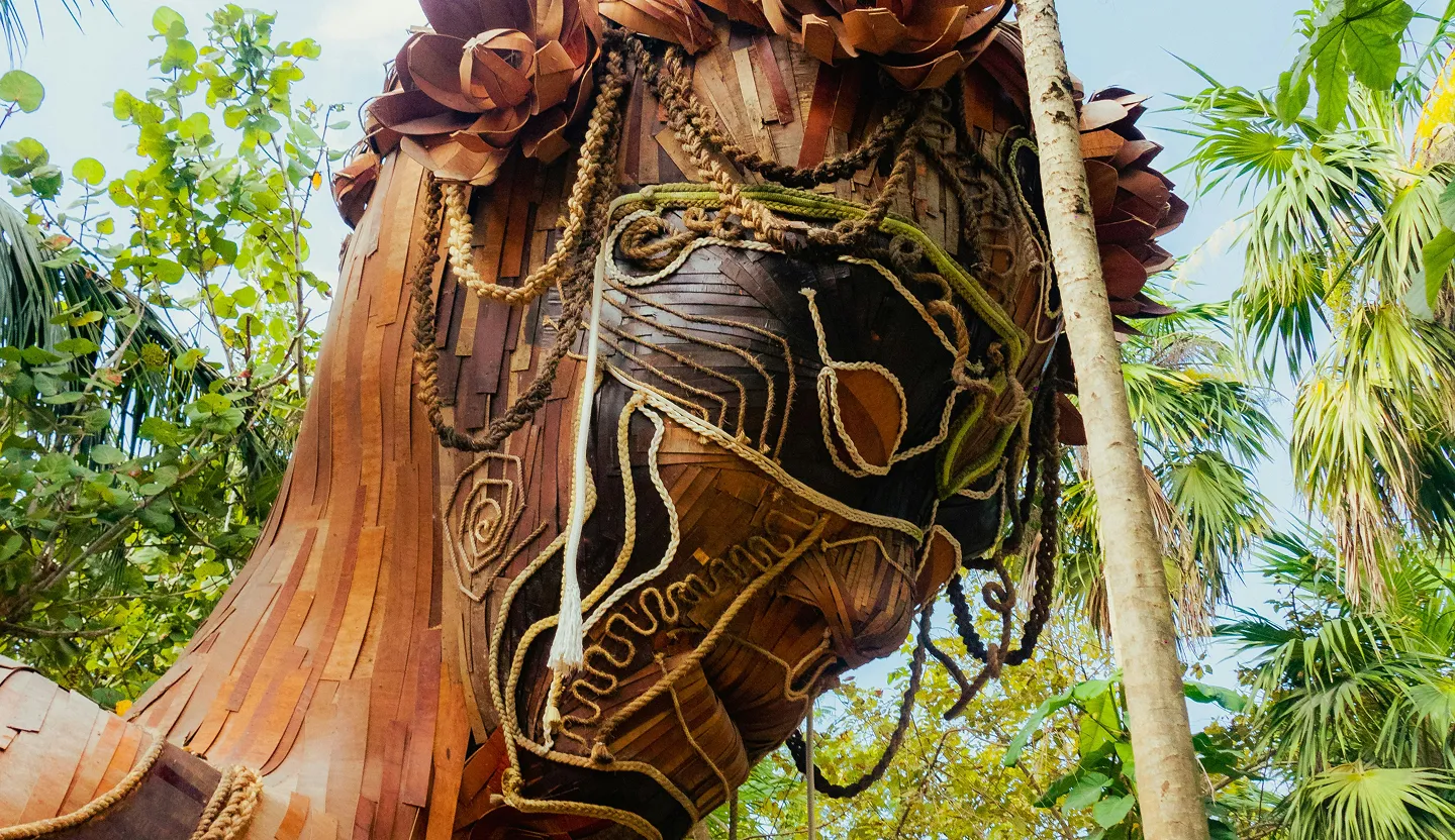 Close-up of a large wooden sculpture of a face adorned with braided ropes, surrounded by lush green tropical plants and a clear sky.