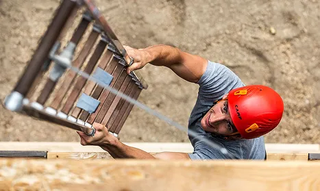 Muž ve šedém triku a červené přilbě šplhá po žebříku opřeném o zdi.