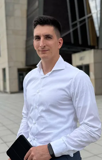 Muž s krátkými tmavými vlasy v bílé košili drží černé pouzdro a má na zápěstí chytré hodinky před moderní budovou.