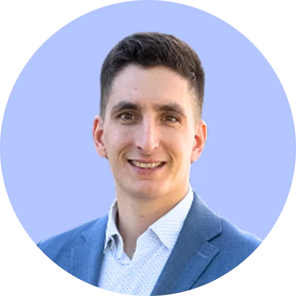 Muž s krátkými tmavými vlasy v modrém obleku a bílé košili se usmívá na světle modrém pozadí.