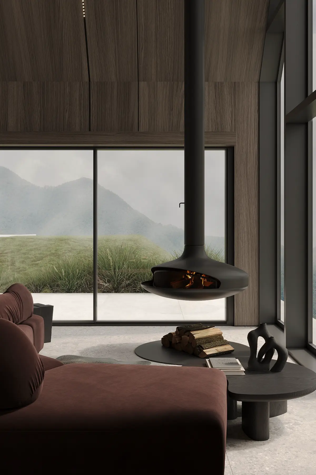 Modern living room with a brown sofa, suspended black fireplace with fire, round black coffee table holding firewood and a black abstract sculpture, large windows showing grassy hills outside.