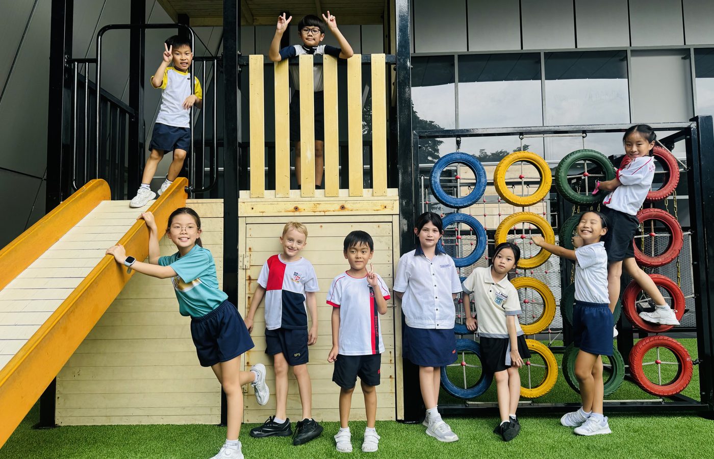 Eight children in their school uniforms climbing on the wooden play frame and tyre wall on artificial grass.