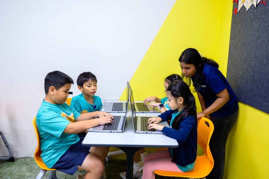 Four children working at laptops around a table while an adult in a navy Agora shirt stands behind them.