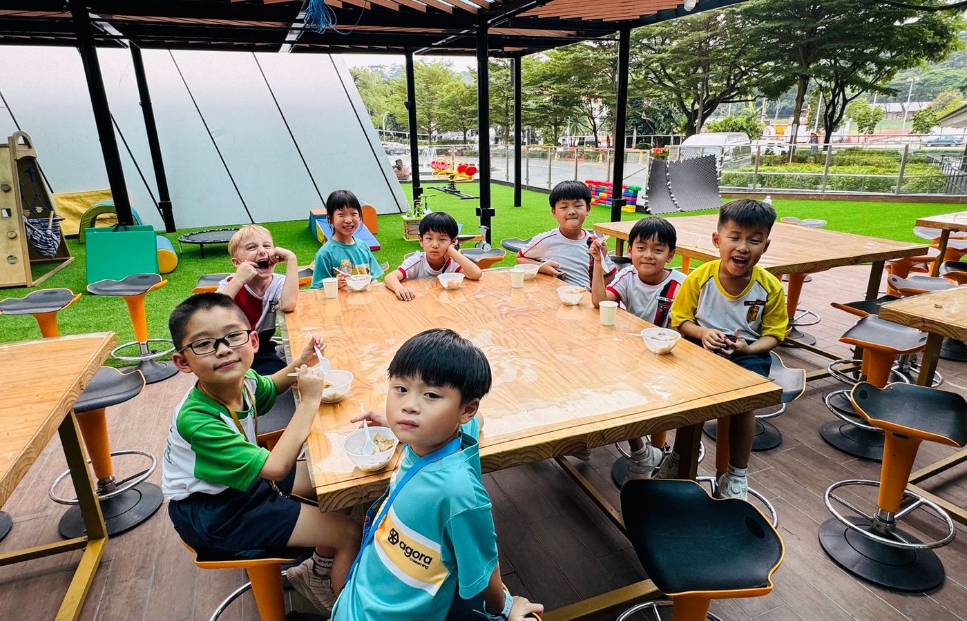 Children in school uniforms and teal Agora polo shirts sitting around a long wooden table on the outdoor terrace, eating from bowls.