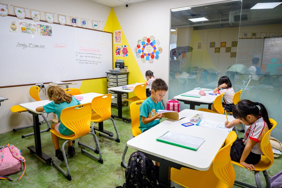 Five children at separate desks in a classroom, each working on their own book, one reading a hardback.