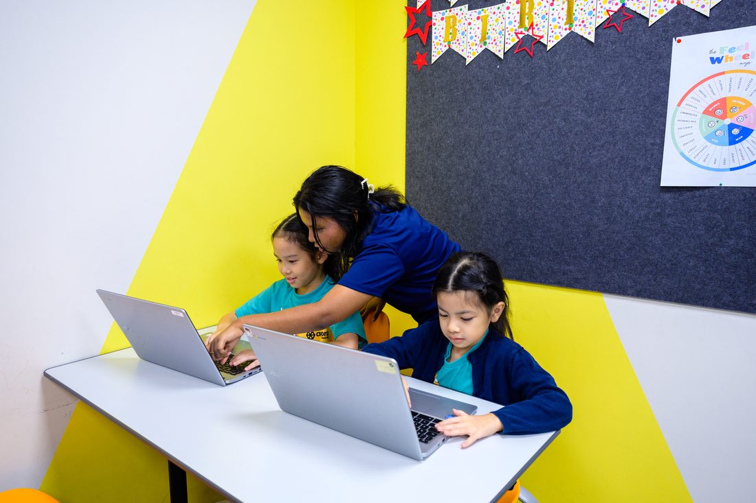 Two girls working at laptops at a white table while an adult in a navy Agora shirt leans in beside one of them.