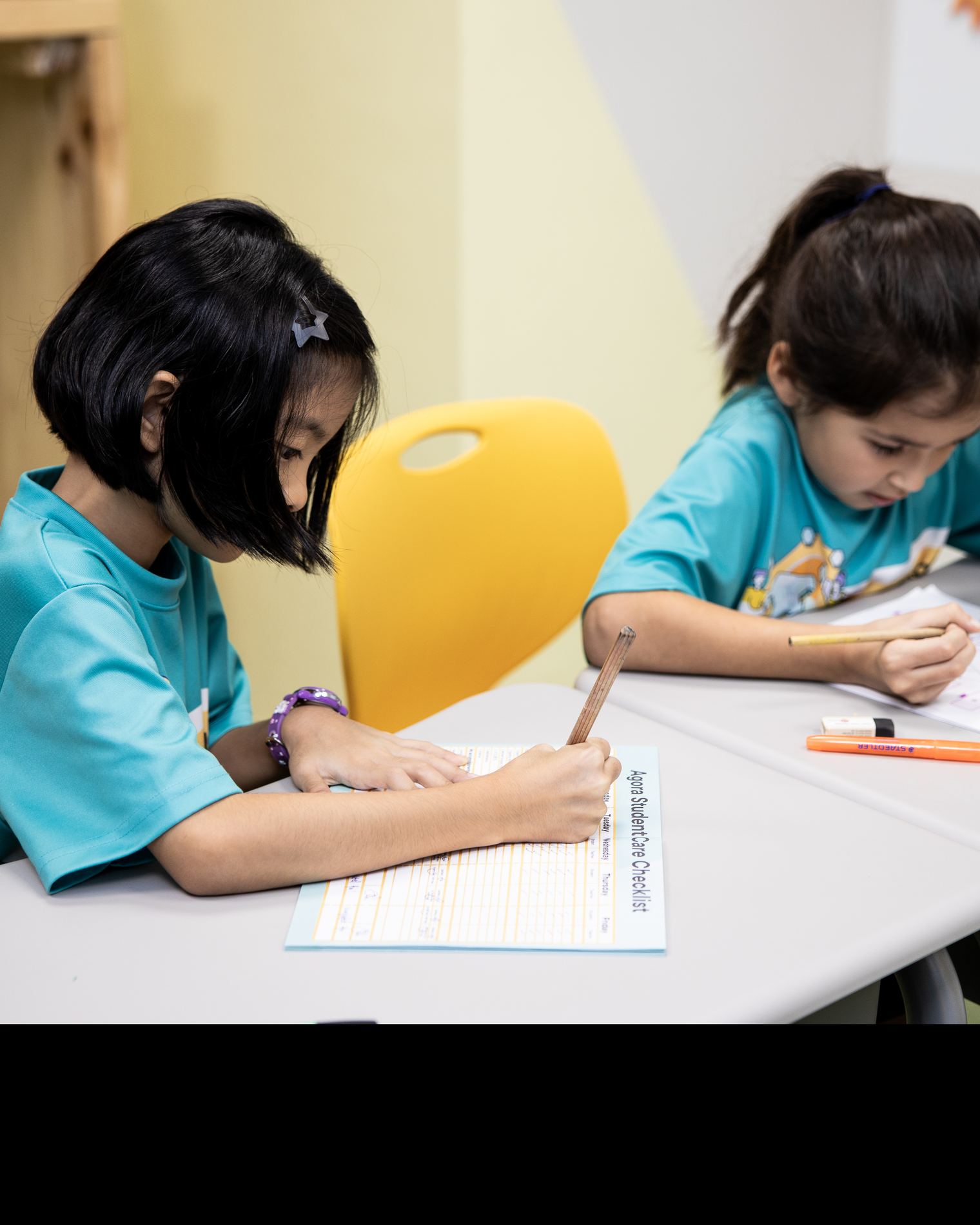 Two children in teal Agora shirts writing at their own desks, both absorbed. The sheet in front reads Agora StudentCare Checklist.