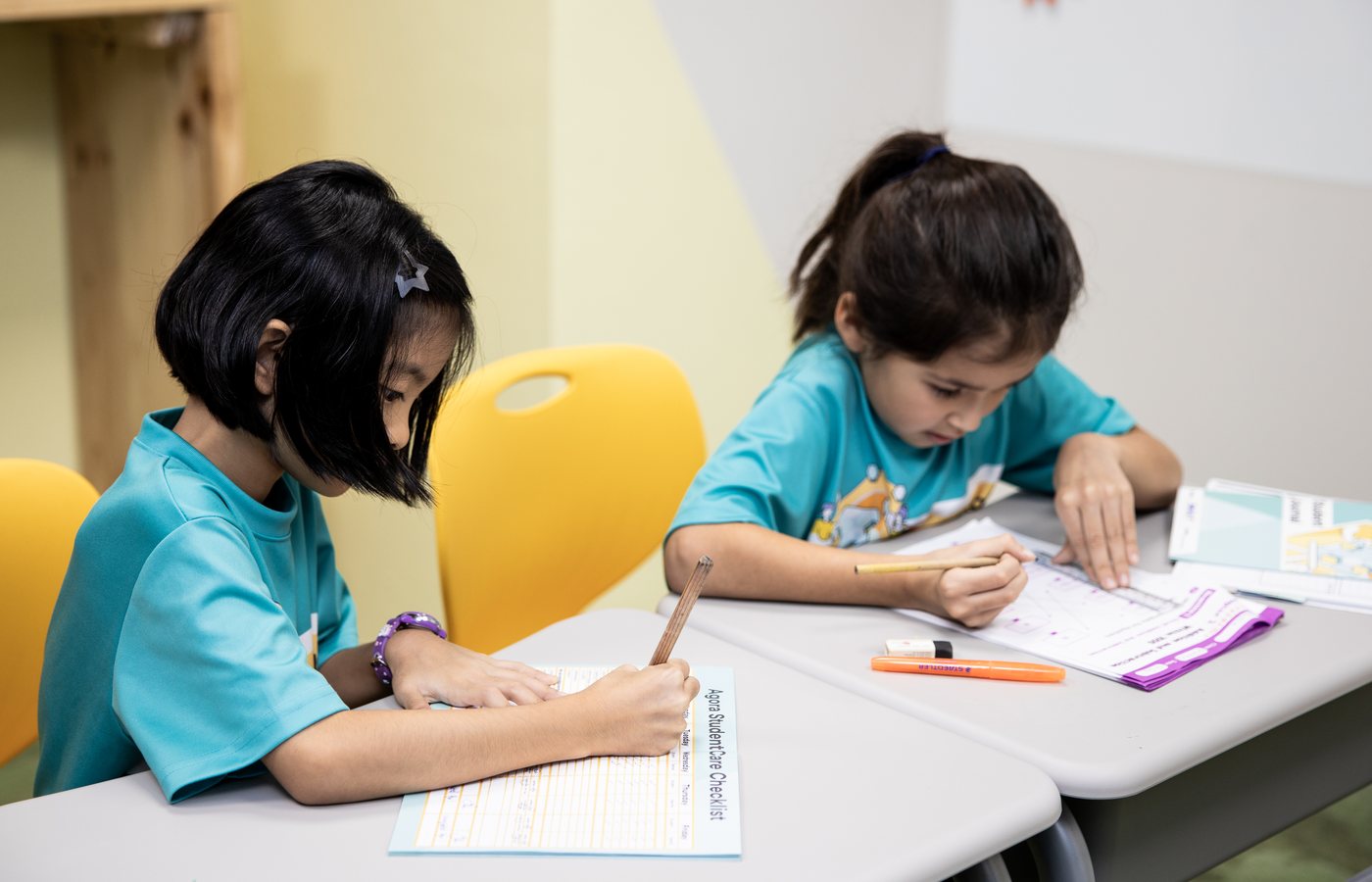 Two children in teal Agora shirts writing at their own desks, both absorbed. The sheet in front reads Agora StudentCare Checklist.