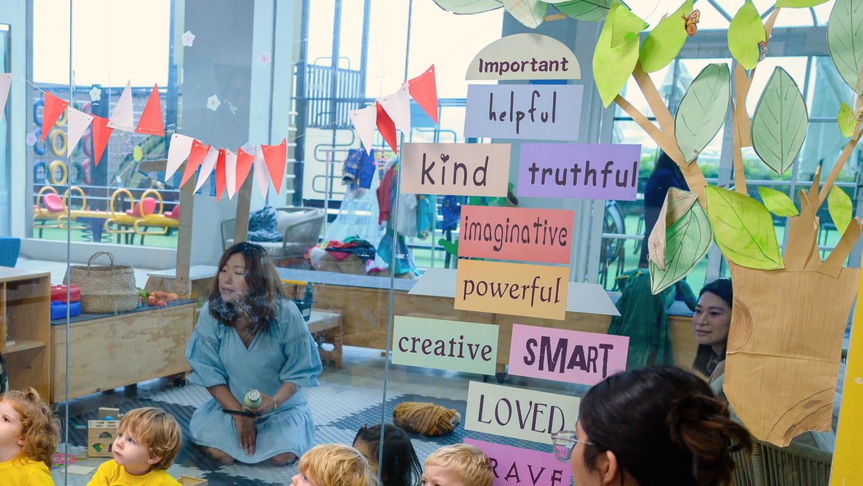 An educator holds up a card to a group of children sitting on the floor, beside a wall of cards reading important, helpful, kind, truthful, imaginative, powerful, creative, smart, loved, brave.