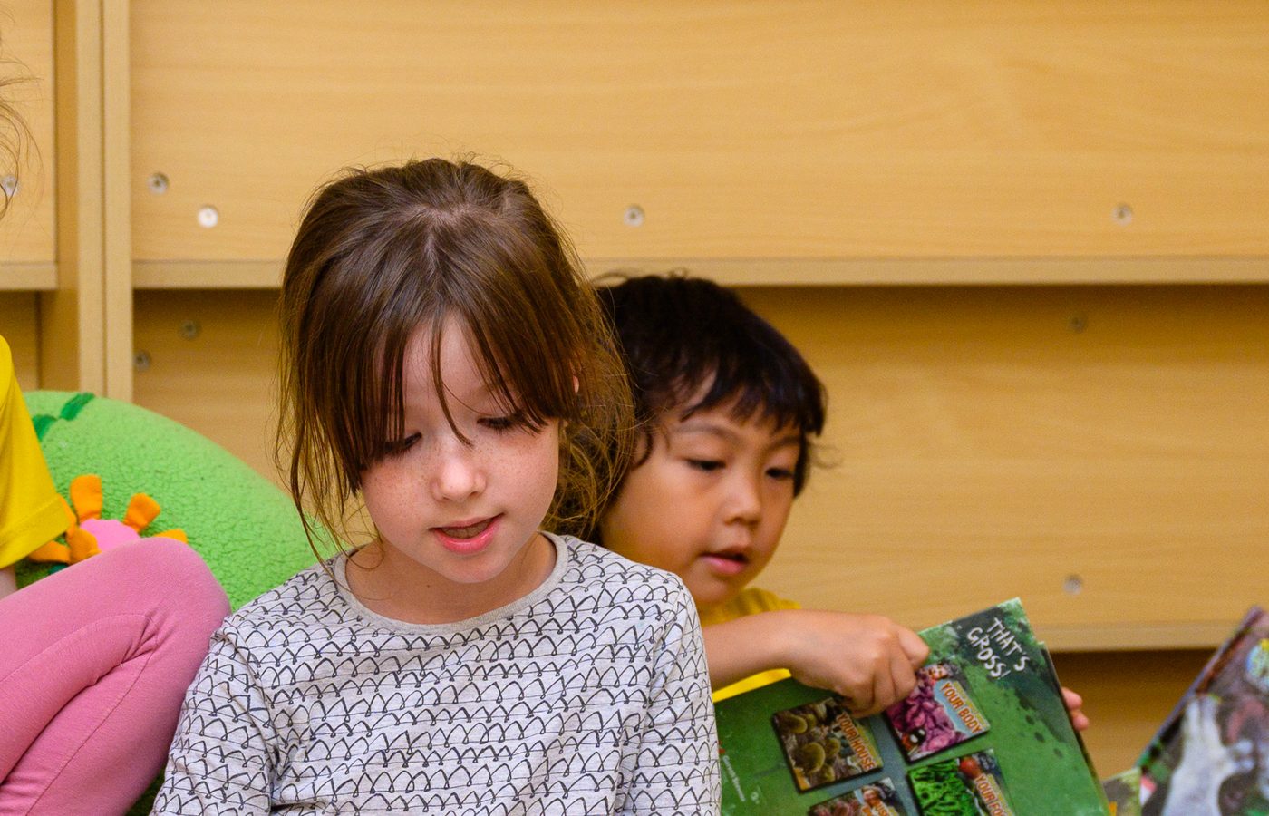 A preschool girl sitting on a beanbag absorbed in an open picture book, while a boy behind her reaches for the one he wants.