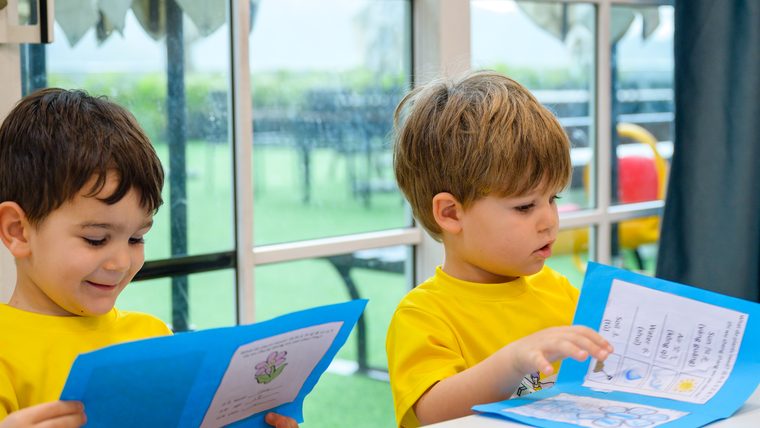 Two boys at a table working on their own printed sheets, one holding a folded blue card open.