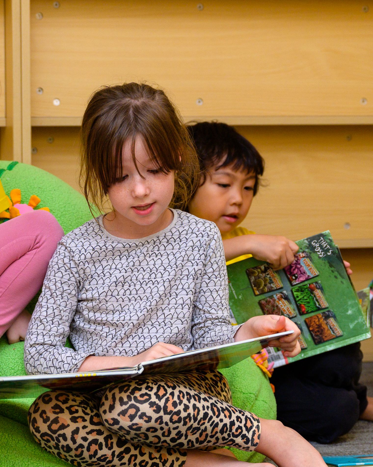 A preschool girl sitting on a beanbag absorbed in an open picture book, while a boy behind her reaches for the one he wants.