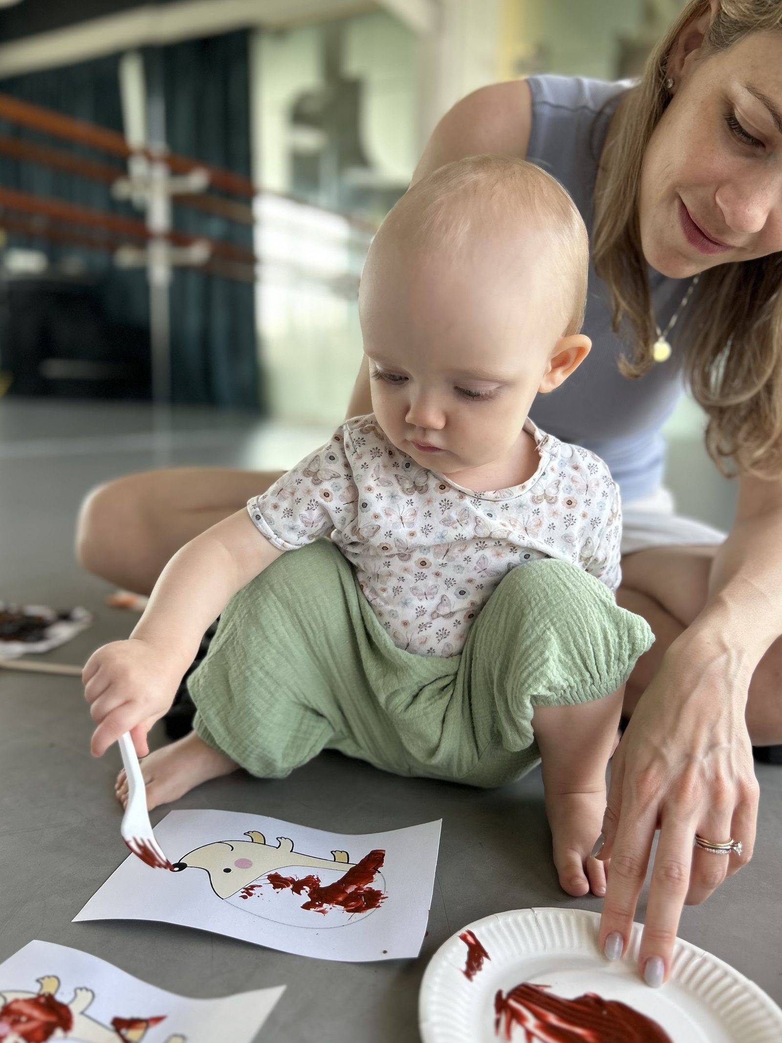 A baby sitting on the floor painting with a fork, an adult kneeling close beside with one hand resting on the floor.