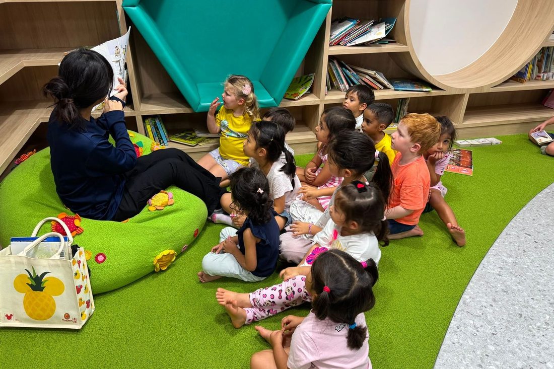 An educator reading to a circle of toddlers on the grass floor of the new early years room, beside curved wooden bookshelves.