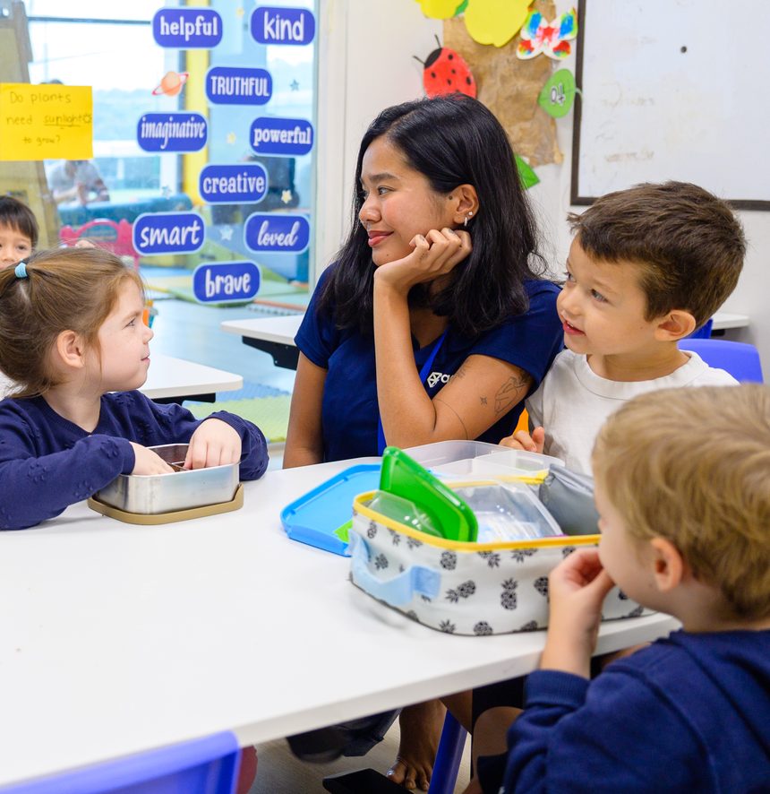 An educator leaning in at a snack table, smiling with a group of children eating from their lunch boxes.