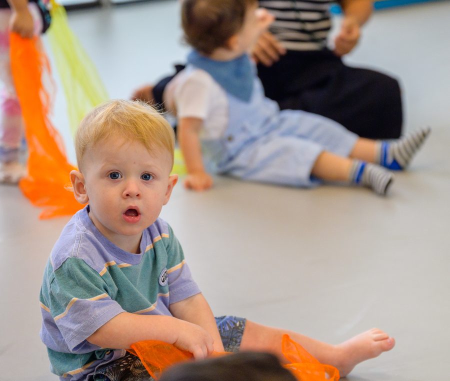 A young child sitting on the floor among loose orange and yellow chiffon scarves, looking straight at the camera.
