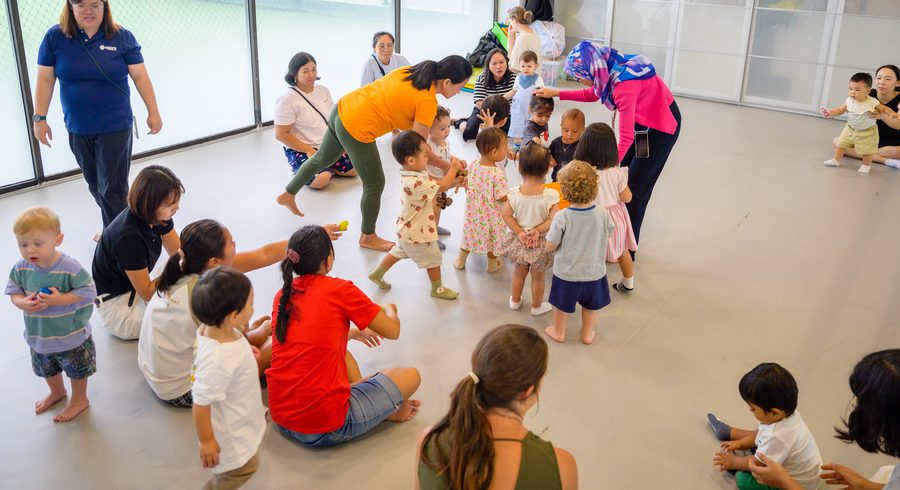 A movement session in a large room: toddlers and their grown-ups together on the floor, with a facilitator in an orange top leaning in among them.