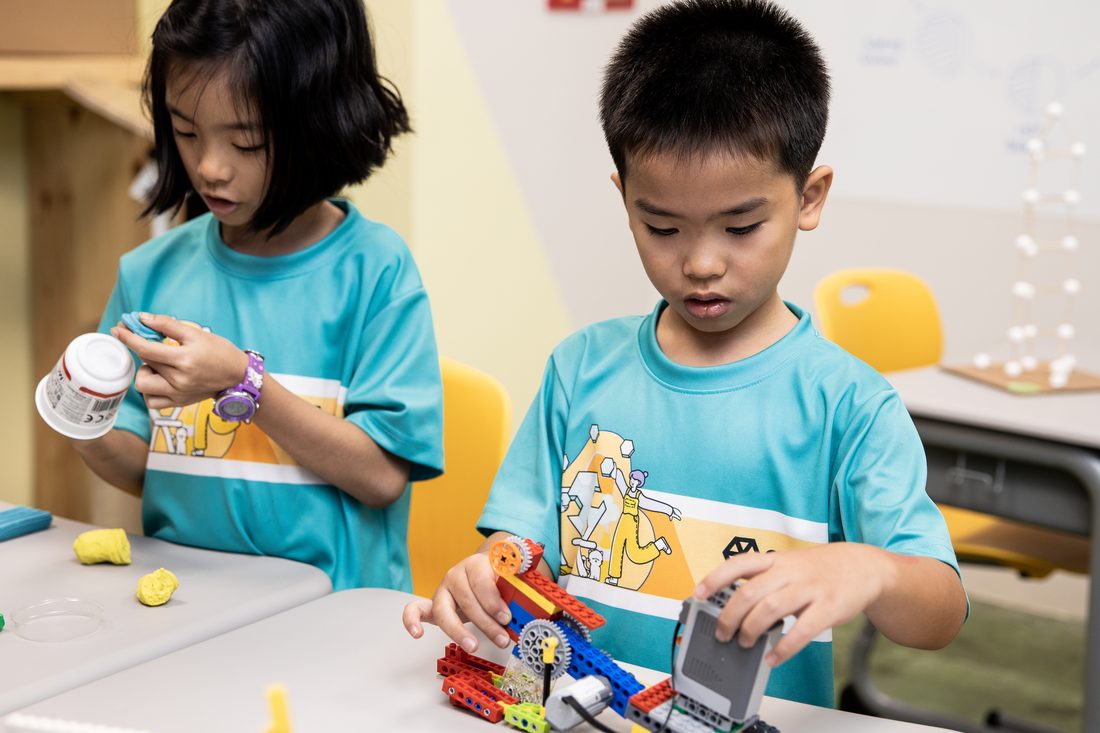 Two children at a table in teal Agora shirts, one opening a pot of modelling clay, the other assembling a geared construction kit.
