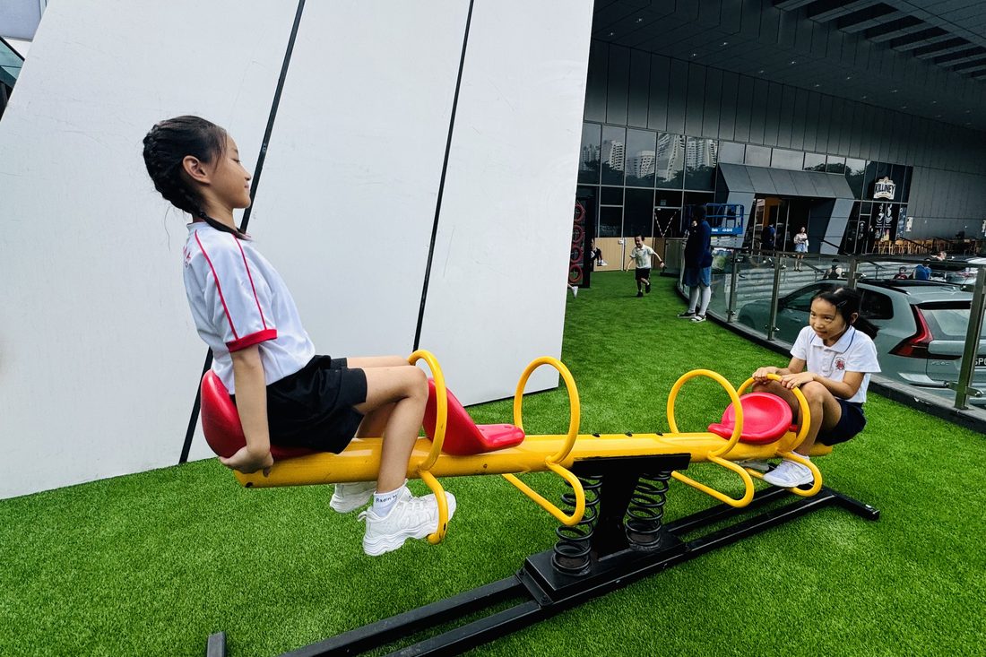 Two children in their primary school uniforms balanced on a yellow seesaw on artificial grass outside, with a glass building behind them.