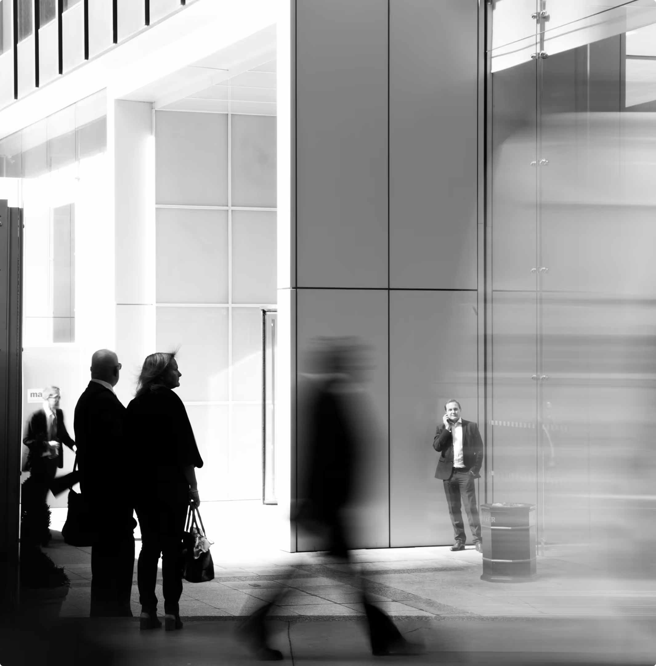 Black and white photo of people in business attire walking and standing near a modern building with glass and metal panels.