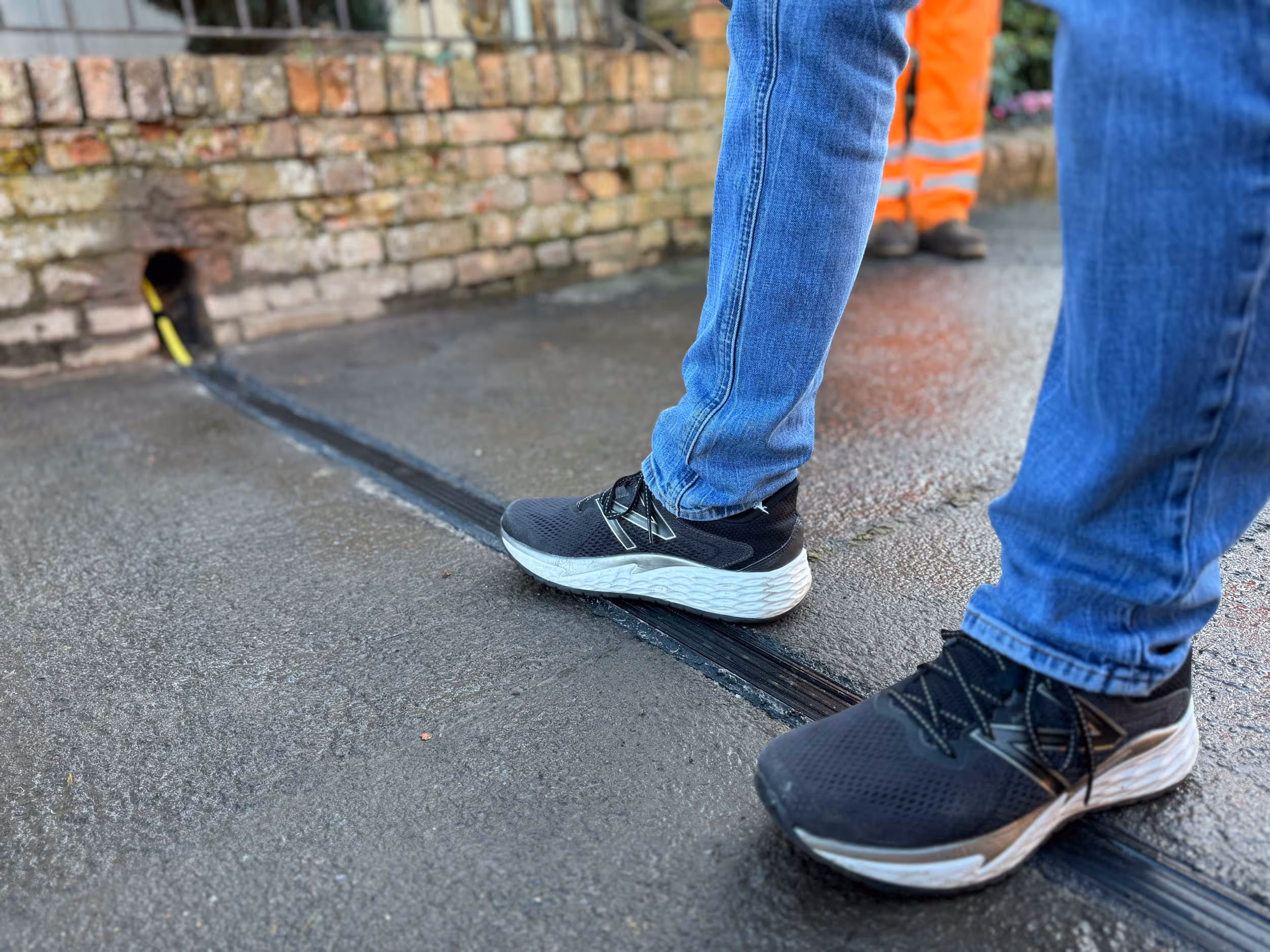 Person wearing black sneakers steps over a cable on a wet street next to a brick wall.