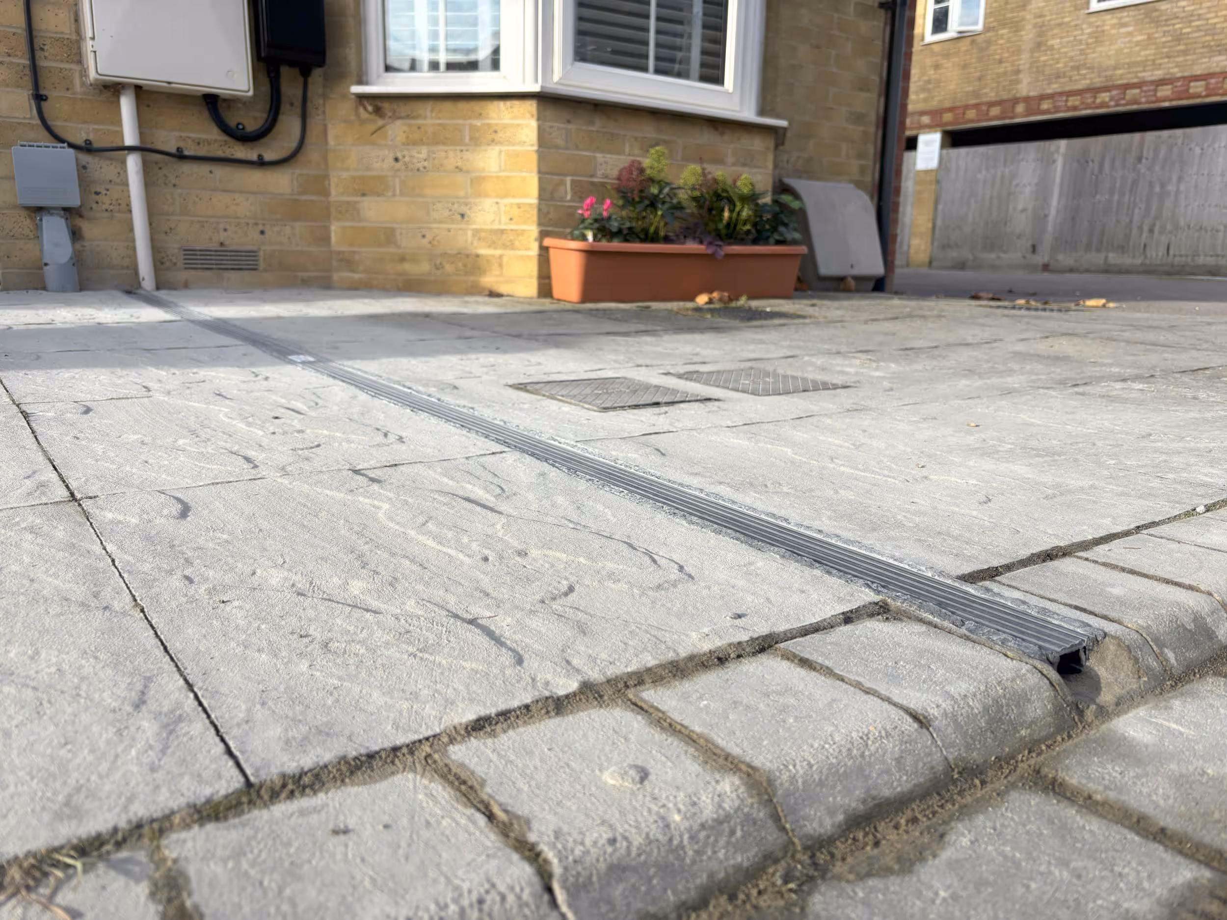 A narrow drainage channel runs across a paved driveway next to a house with a small flower box.