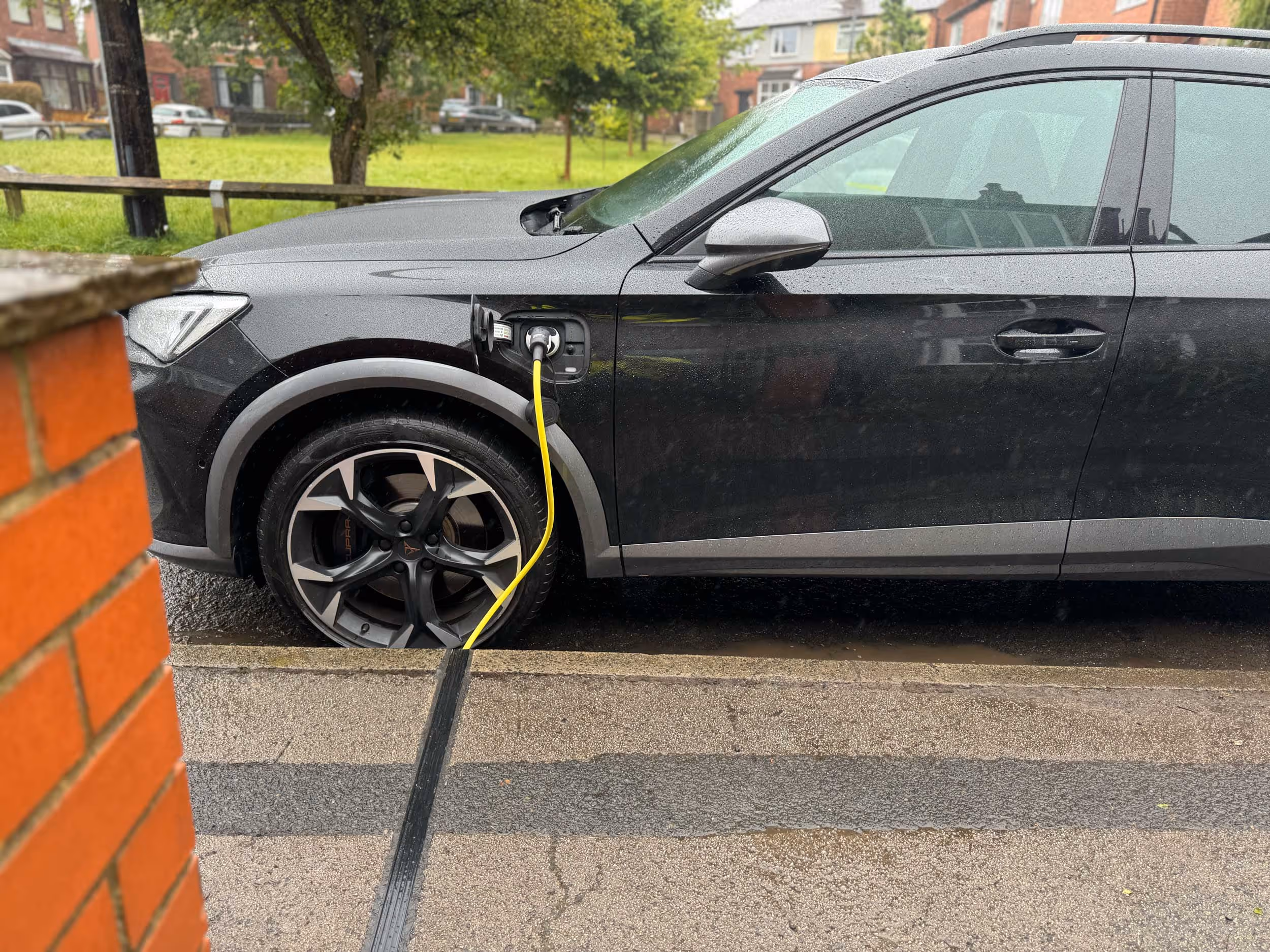 A black car is parked and charging with a yellow cable on a rainy day.