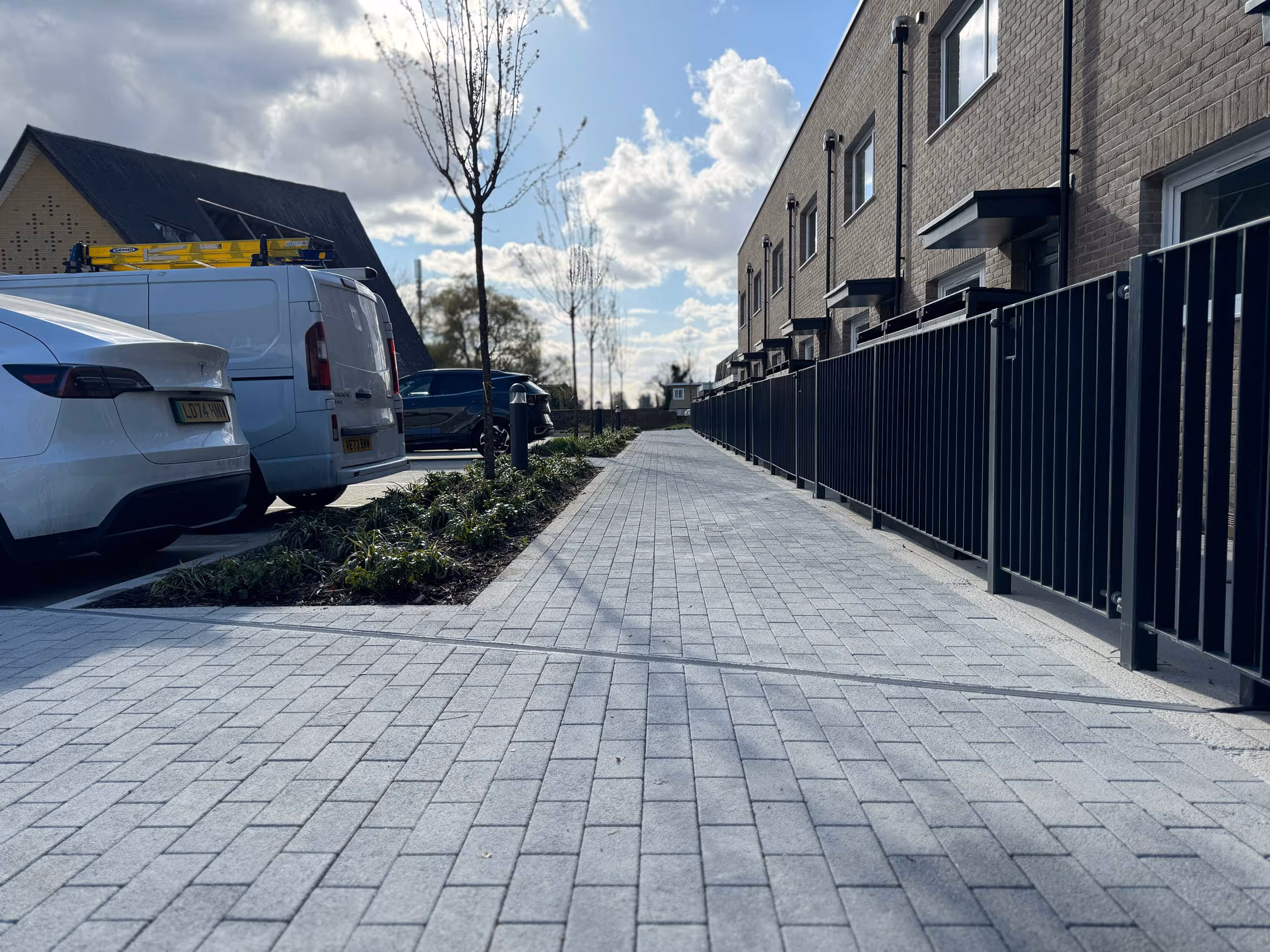 A quiet paved walkway beside parked cars and a row of modern houses on a sunny day.