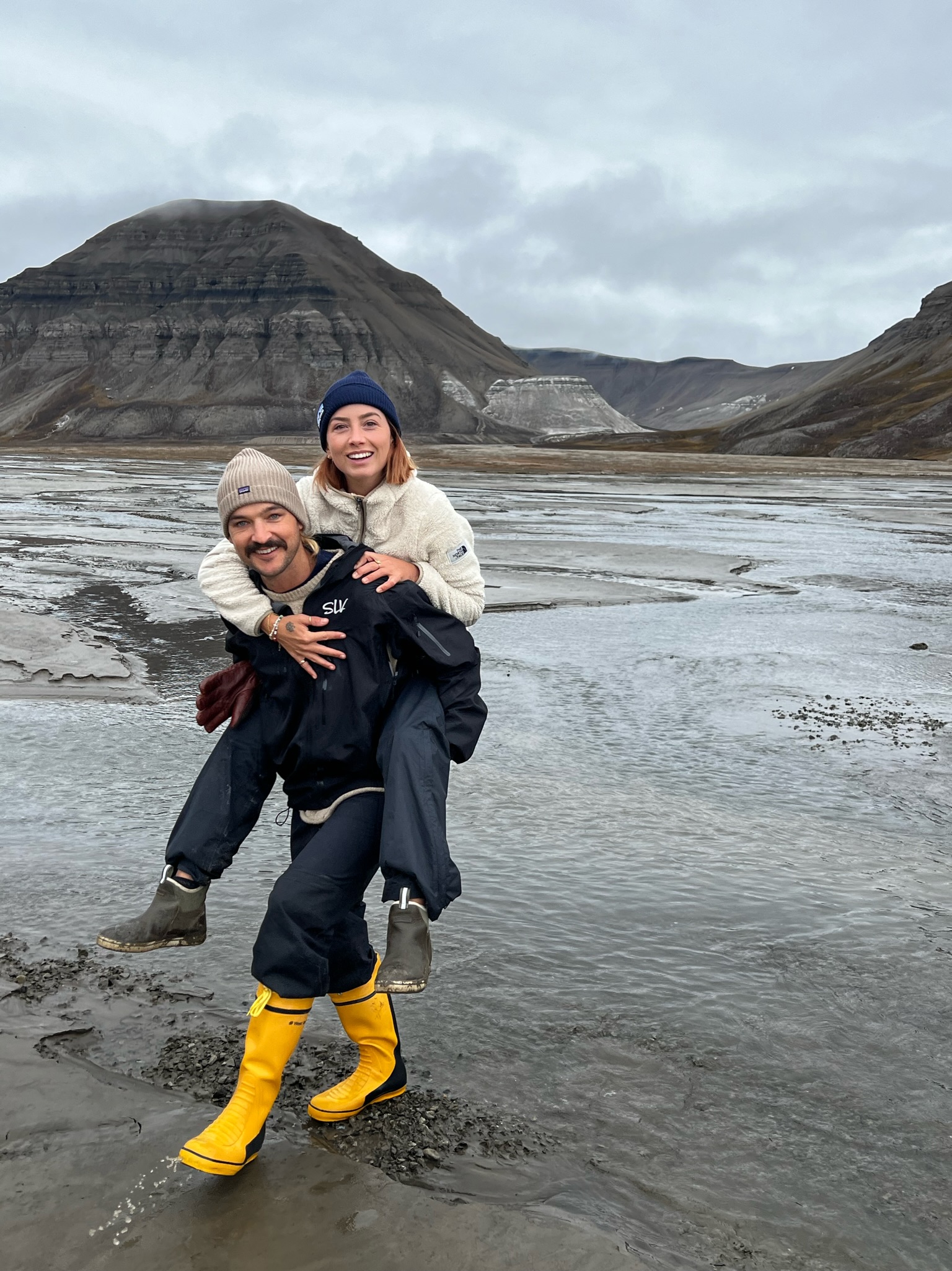 Man wearing bright yellow boots carrying smiling woman on his back near a muddy river with mountains in the background under a cloudy sky.