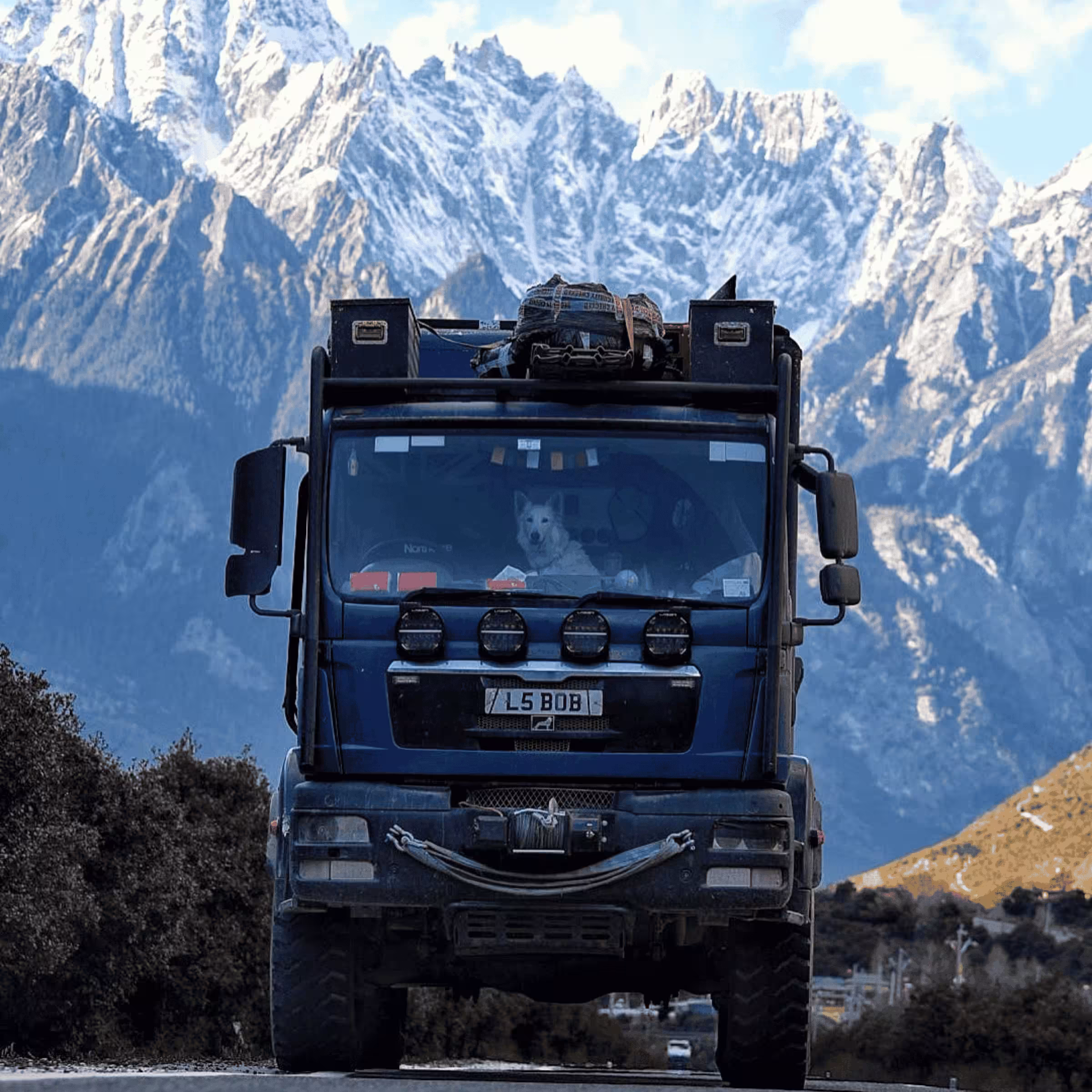 Front view of a blue truck with luggage on top and a white dog inside, set against a backdrop of snow-covered mountains.