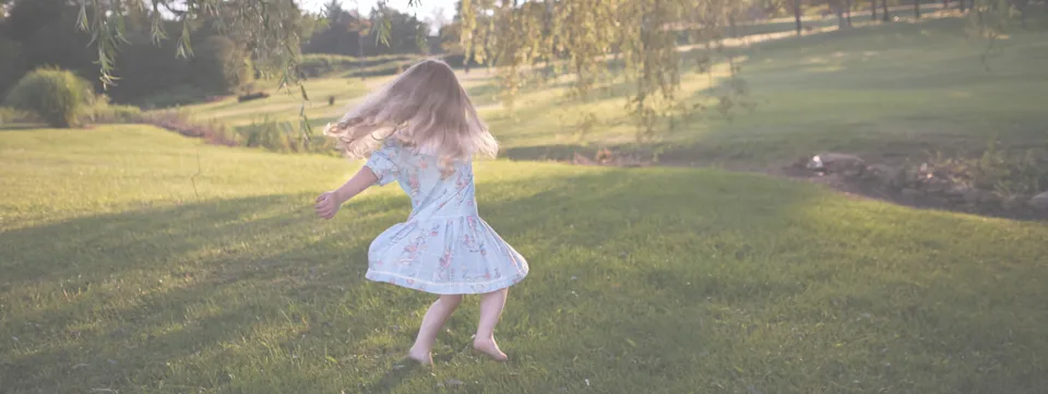 Child with long hair wearing a light blue dress spinning barefoot on green grass in a sunlit park.