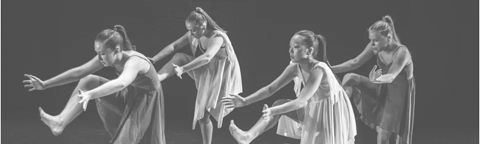 Four female dancers in flowing dresses balancing on one leg with arms extended in a rehearsed pose against a dark background.