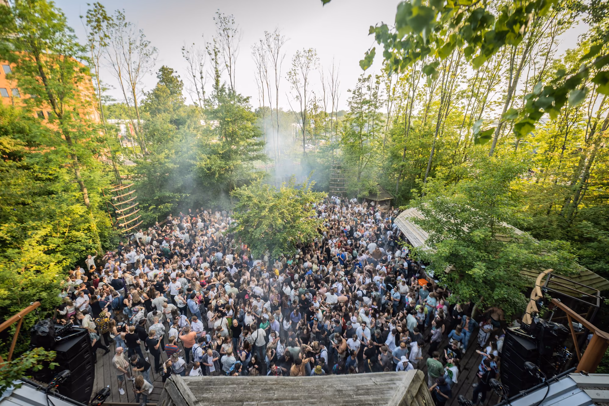 Large crowd gathered outdoors amid green trees, enjoying a daytime event with smoke in the air.