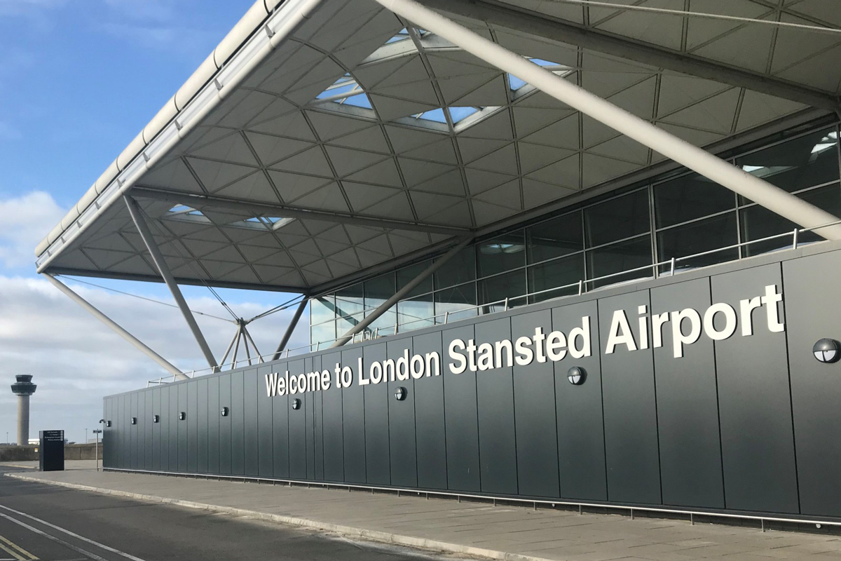 Exterior view of London Stansted Airport with a large sign reading 'Welcome to London Stansted Airport' on a grey building under a modern roof structure.