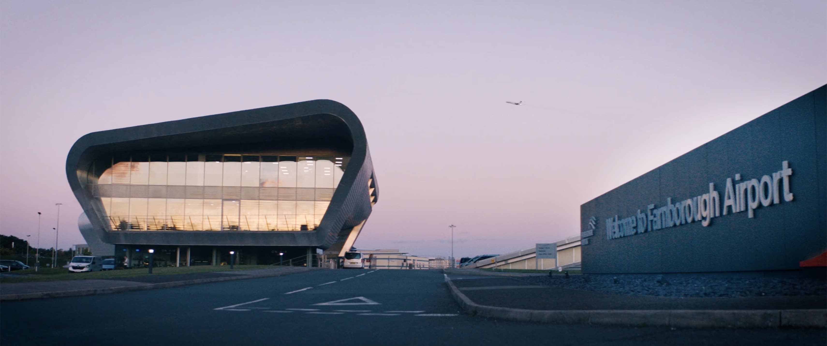 Modern curved building with large glass windows at Farnborough Airport under a twilight sky with an airplane taking off.