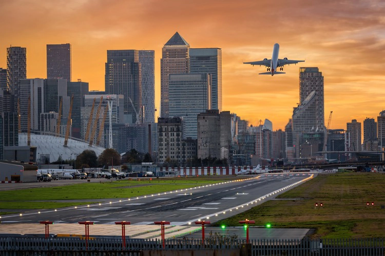Airplane taking off over a runway with city skyscrapers in the background at sunset.