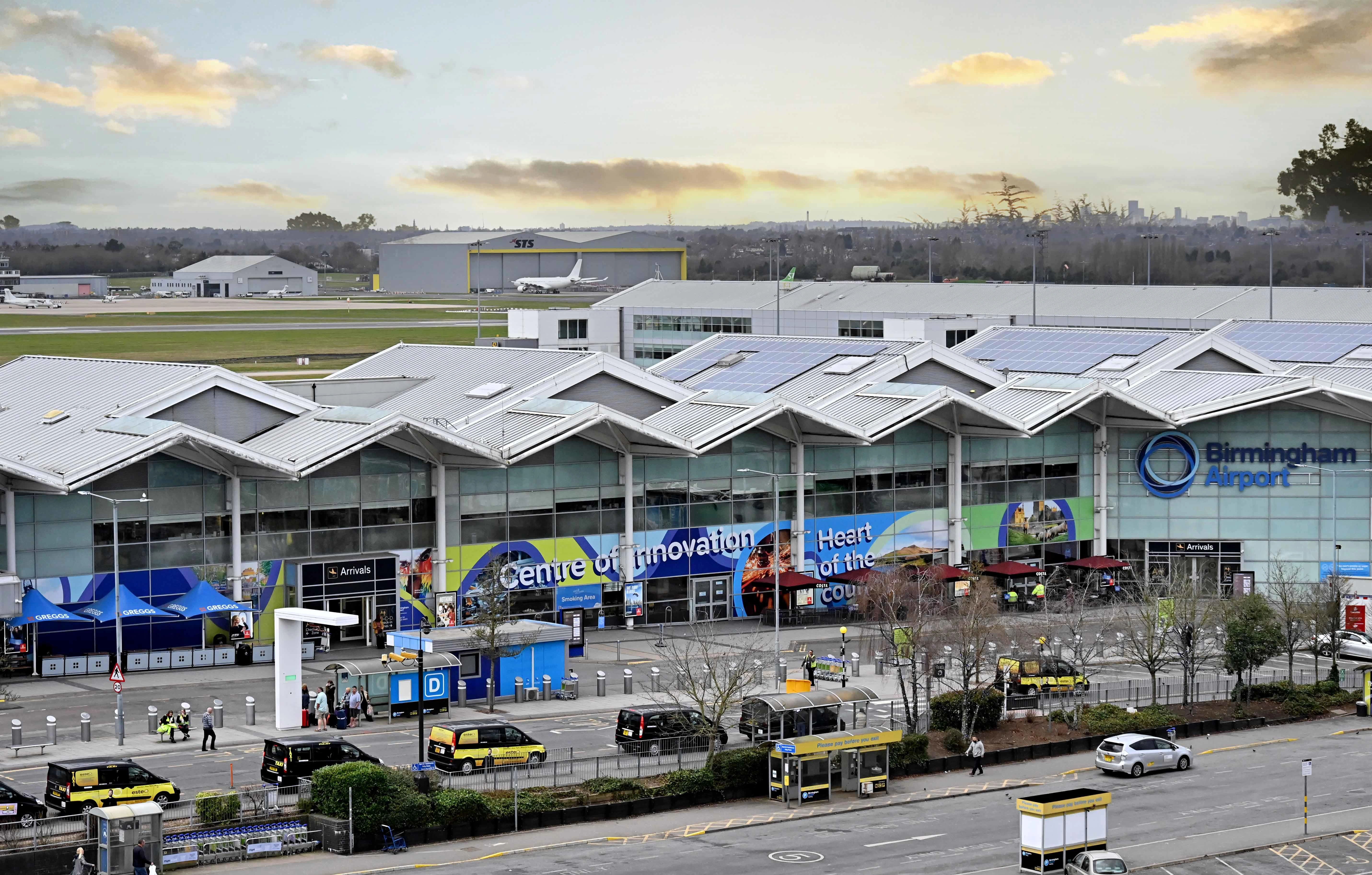 Exterior view of Birmingham Airport terminal with cars parked outside and a cloudy sky.