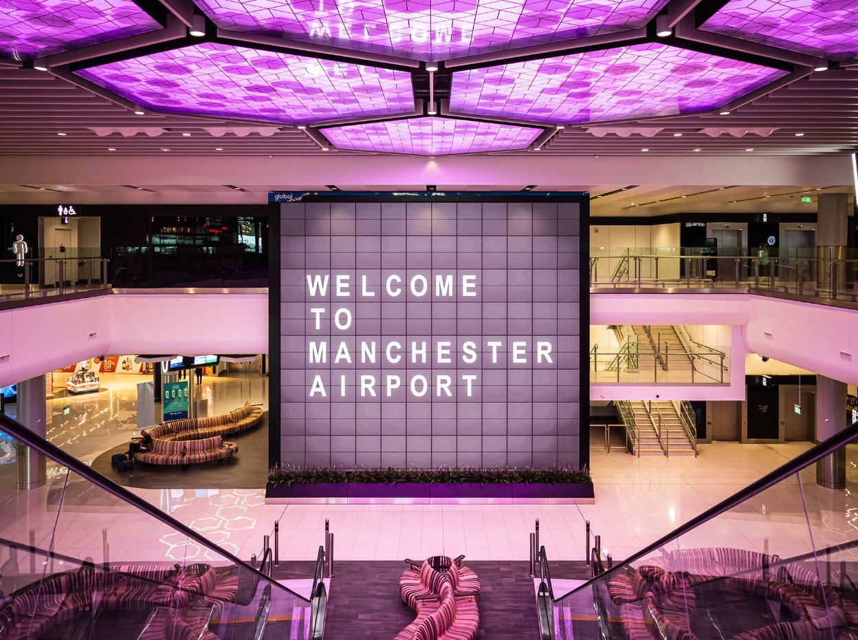 Interior of Manchester Airport featuring a large lit sign reading 'Welcome to Manchester Airport' with purple lighting overhead and modern seating.