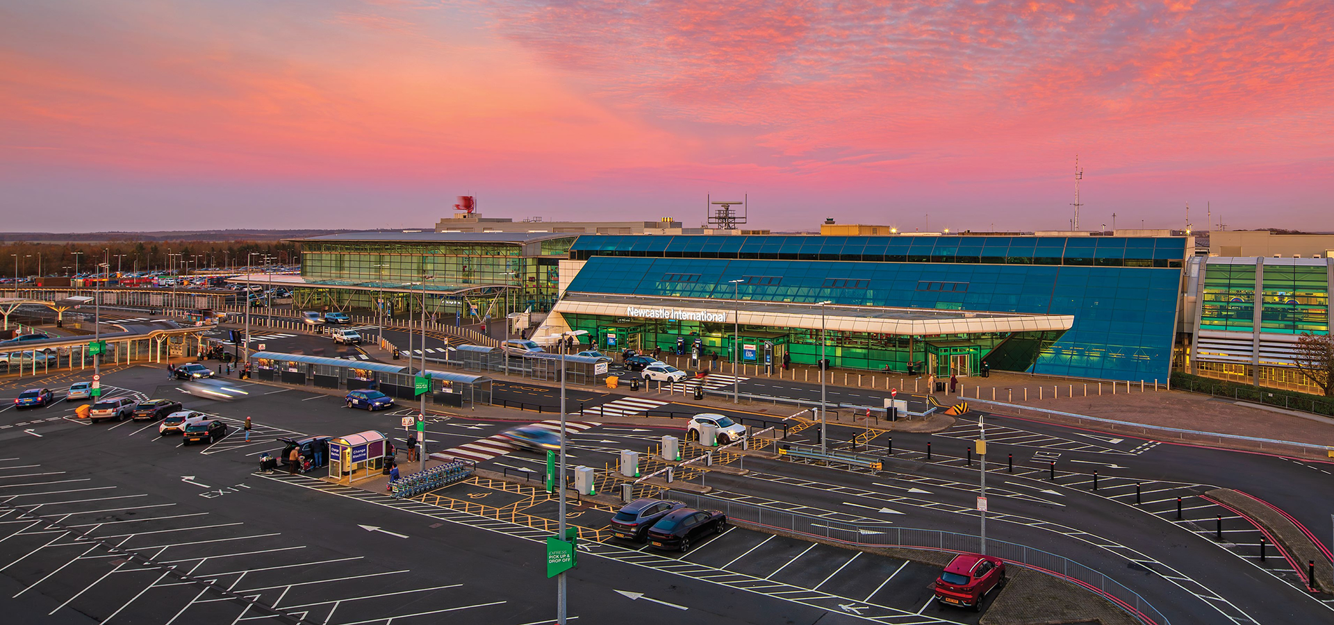 Exterior view of Newcastle International Airport terminal at sunset with parked cars and drop-off zones.