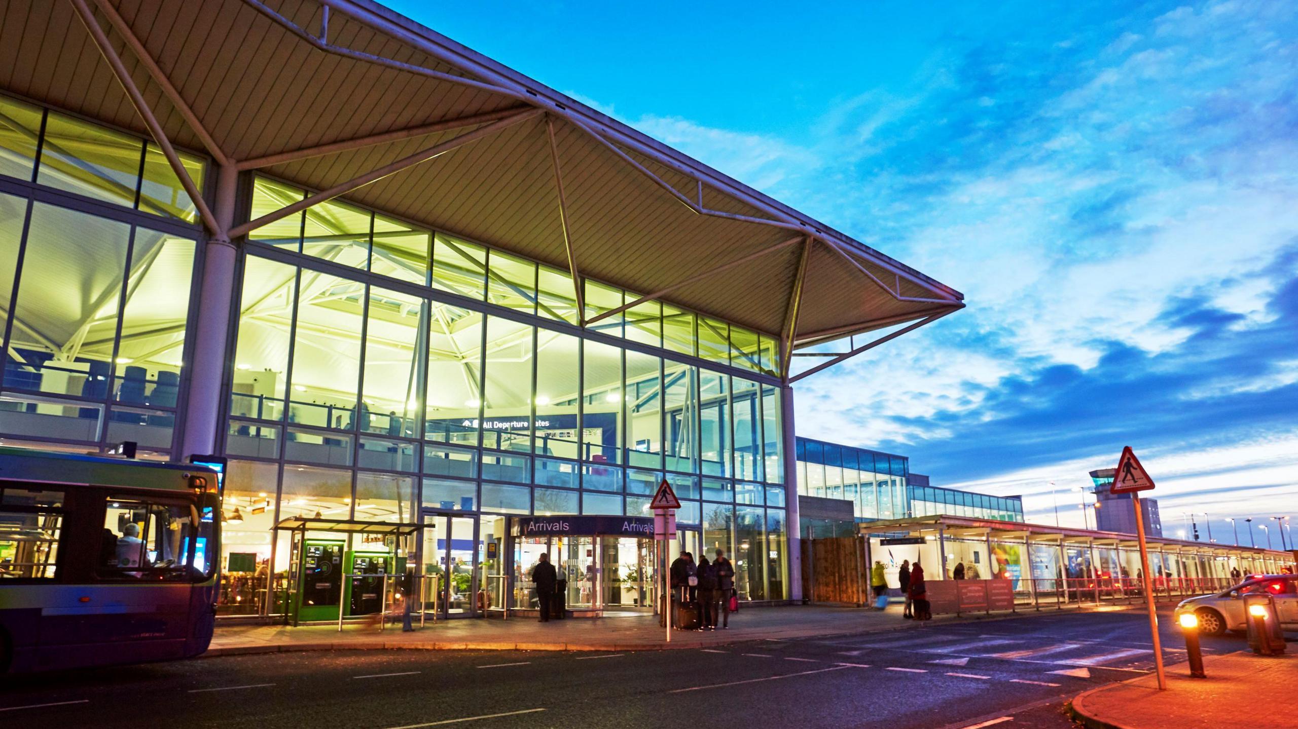 Exterior view of an airport terminal at dusk with large glass windows, people standing outside under an arrivals sign, and a bus on the left.