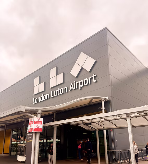 Exterior of London Luton Airport building with people walking near the entrance under a covered walkway.