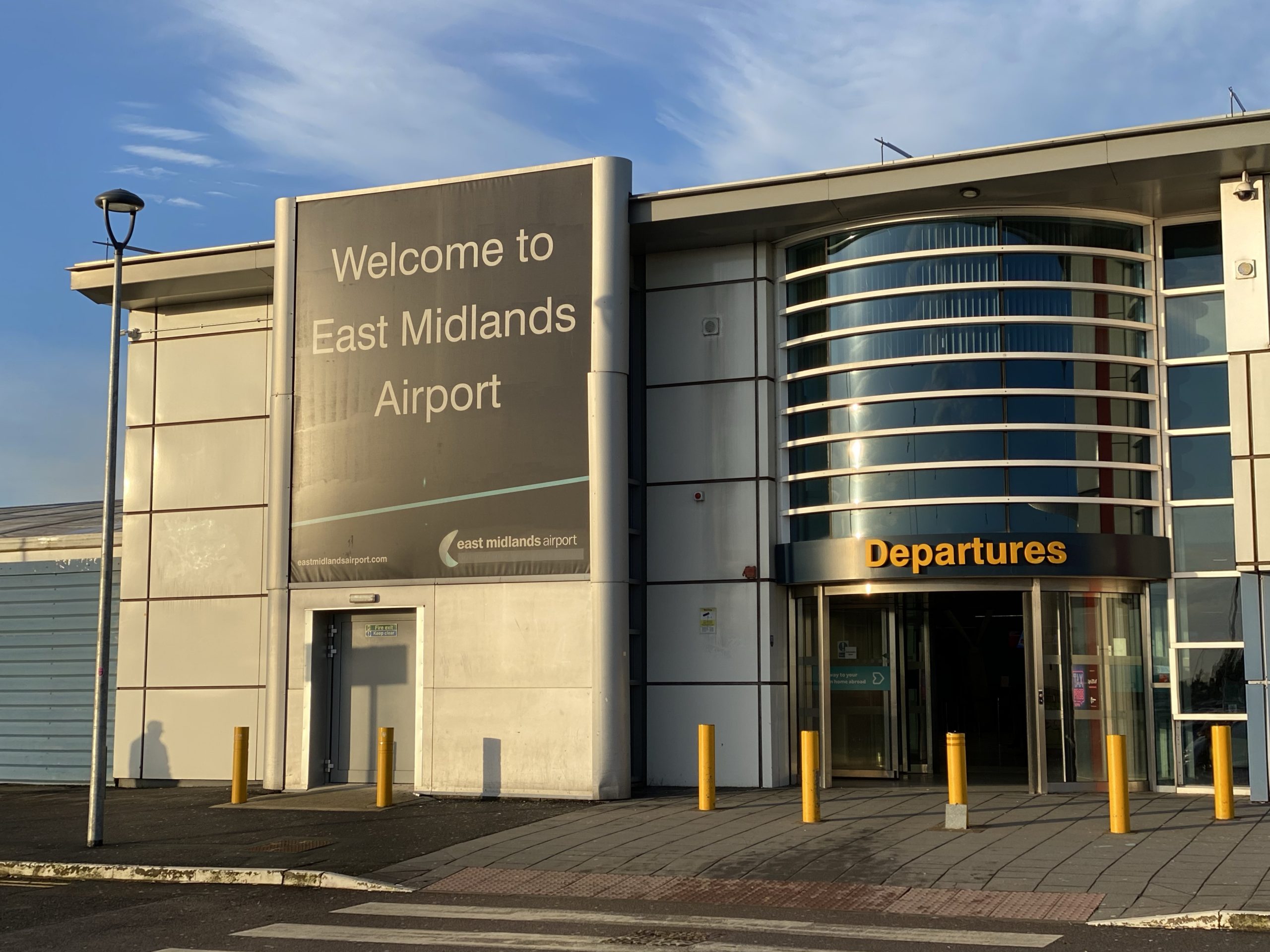 Main entrance of East Midlands Airport showing a large sign that says 'Welcome to East Midlands Airport' and an adjacent entrance labeled 'Departures'.