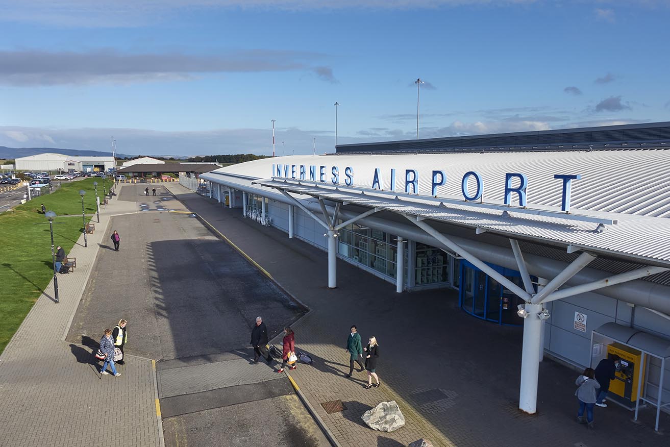 Exterior view of Inverness Airport terminal with passengers walking outside on a sunny day.