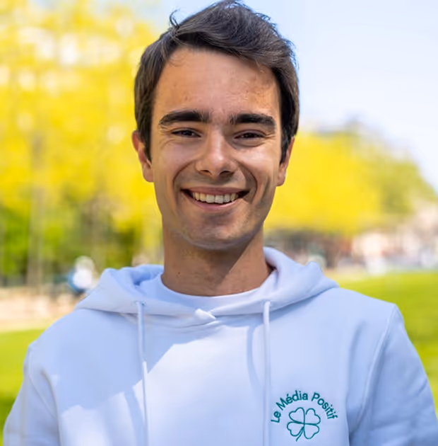 Jeune homme souriant portant un sweat à capuche blanc avec le texte « Le Média Positif » et un trèfle vert, en extérieur avec un arrière-plan flou de verdure.
