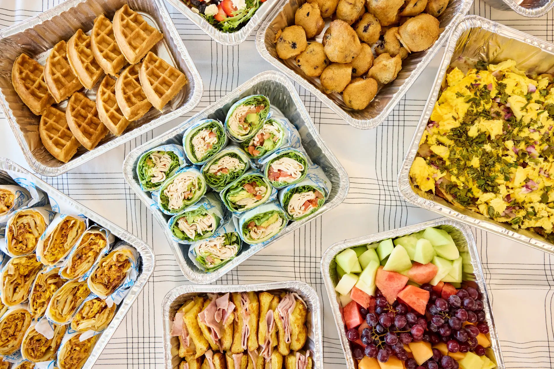 Aluminum trays filled with breakfast foods including waffles, fruit salad with grapes and melon, scrambled eggs with herbs, ham sandwiches, wraps with lettuce and turkey, muffins, and breakfast burritos.