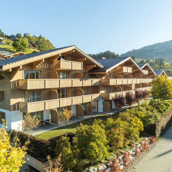 Mehrstöckige Holzchalets mit Balkonen, umgeben von grünen Büschen und Bäumen in einer sonnigen Berglandschaft.